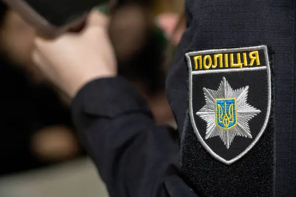 Koyu renk üniformalı bir polis memuru belirgin bir rozetle ayakta duruyor. Bu durum, büyük olasılıkla halka açık bir toplantıda veya etkinlikte yasa uygulama görevlerinde yer aldığını göstermektedir..