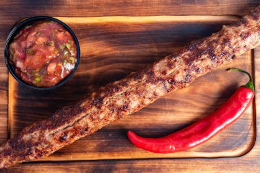 Uzun ızgara bir kebap küçük bir kase taze salsa sosunun yanında ahşap bir tahtanın üzerinde duruyor. Kırmızı biber çok canlı bir renk katar ve çok cazip bir aşçılık sunumu yaratır..