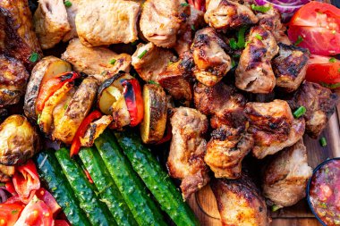 Canlı bir ızgara et ve ahşap tabakta dizilmiş renkli sebzeler. Taze domates, salatalık ve şişler iştah açıcı açık hava yemeğine ilave eder..