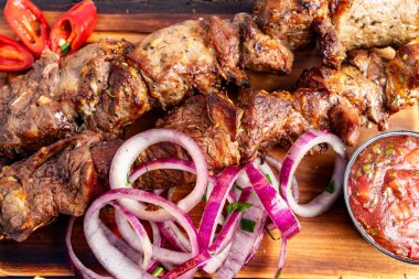 Sulu ızgara et şişleri, taze soğanlar ve küçük bir kase baharatlı salsa sosuyla birlikte ahşap bir tabakta sergileniyor. Renkli kırmızı biberler biraz canlılık katar..