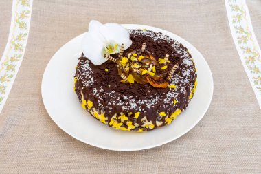 Üzerinde orkide ve çikolata parçacıkları olan çok güzel sunulmuş bir çikolatalı pasta beyaz bir tabakta duruyor. Tatlı desenli bir masa örtüsüne konur ve dikkat çeker..