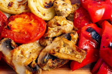 Ahşap tabakta domates, soğan ve biber parçaları içeren renkli ızgara sebzeler, açık havada canlı bir yemek deneyimi sergiliyorlar..