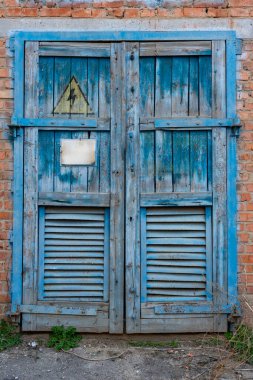 Ahşap çatallı sıkıntılı mavi kapılar endüstriyel bir ortamda duruyor. Uyarı işaretleri çevredeki elektriksel tehlikeleri, yaşı ve ihmalleri gösteriyor..