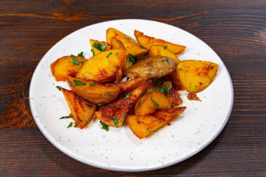 Lezzetli kızarmış patatesler taze yeşil otlarla süslenmiş beyaz bir tabağa saçılmıştır. Koyu ahşap yüzeyde altın rengi patatesler parlıyor.