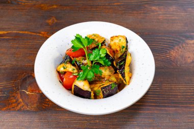 Patlıcan ve domatesleri de içeren canlı bir tabak taze otlarla kaplı, ahşap bir masada yuvarlak beyaz bir kasede sunuluyor.