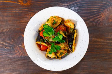 Kızarmış tavuk, patlıcan ve taze maydanozla süslenmiş domatesli renkli bir yemek. Canlı yemek, benekli beyaz bir tabakta ahşap bir arka planda servis edilir.