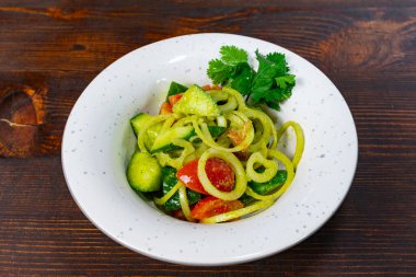 Bu yemek, kişniş soslu salatalık ve taze domateslerin canlı bir karışımını içeriyor. Tahta bir arka planda beyaz bir kasede servis edilmiş. İştah açıcı ve sağlıklı görünüyor.