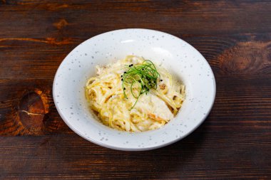 Kremalı bir makarna tabağı, yeşil otlarla süslenmiş basit beyaz bir kâse içinde sunulur. Kırsal ahşap masa, çekici mutfak sunumuna sıcaklık katıyor..