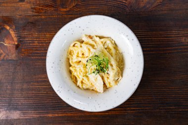 Bir kase kremalı makarna, sade ahşap masa raflarında servis edilir. Üstü şifalı otlarla kaplı erişte. Yemek zengin ve davetkar görünüyor. Tatmin edici bir yemek için mükemmel..