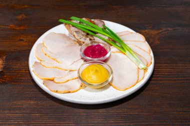 Bir tabakta, sarı sos ve kırmızı sos da dahil olmak üzere çeşitli sosların yanında dilimlenmiş etler yer alıyor. Yeşil soğan garnitürleri sunumu güçlendirir, koyu ahşap bir masaya kurulur.