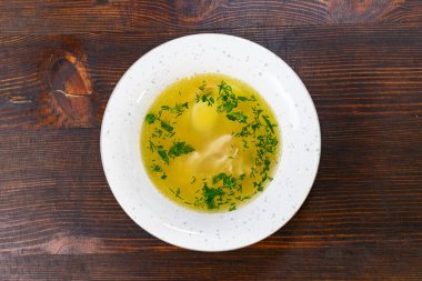 Sıcak bir kase altın çorbada taze et parçaları bulunur ve taze yeşil otlarla süslenir. Tabak, kırsal ahşap bir zemine dayanıyor. Yemeğin sıcaklığını vurguluyor.