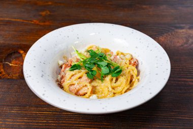 Kırsal bir ahşap masada bir kase kremalı carbonara sunulur. Makarna taze otlar ve serpilmiş peynirle süslenir. Lezzetli bir yemek deneyimi vadeder..