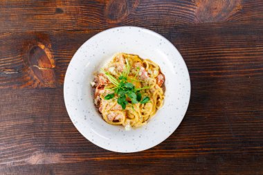 Taze otlar ve rendelenmiş peynirle süslenmiş kremalı bir makarna tabağı benekli beyaz bir tabakta oturur. Kırsal ahşap masa bu iştah açıcı yemek için sıcak ve davetkar bir zemin sağlar..