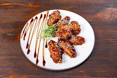 Bir tabak, sosla serpiştirilmiş ve susam tohumları ve mikroyeşilliklerle süslenmiş sulu tavuk kanatları içerir. Ahşap arka plan yemeğin çekiciliğini arttırır..