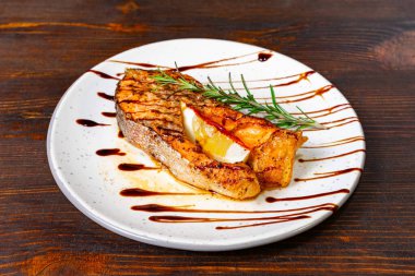 Taze otlar ve limonla süslenmiş balzamik redüksiyonla süslenmiş güzel bir ızgara balık fileto. Kırsal ahşap arka planda beyaz bir tabakta servis edilmiş..