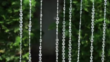 Kristal boncuklar bahçe ortamında pırıl pırıl bir zemin oluşturur. Narin kristal boncuklar dikey ipliklerde asılı, yemyeşil bir bahçede güneş ışığını yansıtıyor. Canlı yeşillik büyülü ortamı güçlendiriyor, sakin bir atmosfer yaratıyor.