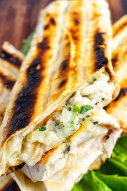 Ahşap bir yüzeyde erimiş peynir ve taze otlarla dolu ızgara yassı ekmek yığını sergileniyor. Çıtır çıtır dış görünüş yumuşak, yapışkan iç güzellikle zıtlaşır ve ağız sulandırıcı bir zevk verir..