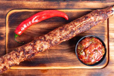Uzun, ızgara bir kebap, bir kase baharatlı salsa ve taze kırmızı biberle birlikte ahşap bir tahtanın üzerinde duruyor. Sunum iştah açıcı dokuları ve canlı renkleri vurguluyor.