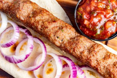 Lezzetli bir kebap taze kırmızı soğanla süslenmiş ve bir fincan salsa sosuyla servis edilir. Bu yemek zengin dokuları ve canlı renkleri vurgular.