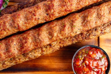 Sulu baharatlı et şişleri ahşap bir tahtanın üzerinde sunuluyor. Yanında bir kase taze salsa, canlı renkler ve bir ziyafet için uygun dokular sergileniyor..