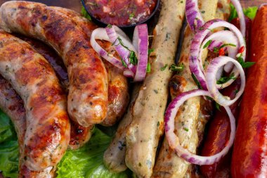 Bir tabakta çeşitli ızgara sosisler düzenlenir, yanında da kırmızı soğan dilimleri ve bir kase taze salsa sosu bulunur. Canlı renkler lezzetli bir yemek için iştahı davet eder..