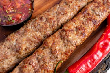 İki sulu ızgara kebap, bir kase baharatlı salsa sosu ve taze kırmızı biberle birlikte ahşap tabakta dinleniyor. Tabakta zengin lezzet ve canlı renkler sergileniyor..