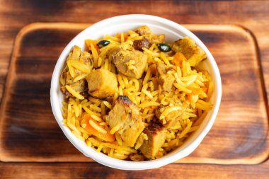 Lezzetli sarı pirinçle dolu beyaz bir kase yumuşak tavuk parçaları ve renkli sebzelerle ahşap bir yüzeyde duruyor. Bu yemek lezzetli bir yemek deneyimini davet ediyor..