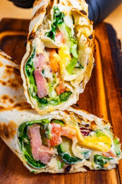 Tahtaya yeni bir naylon konur. Renkli yeşillik, et ve peynir katmanları ortaya çıkar. Paket kusursuzca kızartılmış, iştah açıcı dokusunu ve lezzetlerini gösteriyor..