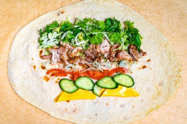 Taze pişmiş et, bir mutfak ortamında yumuşak tortilla üzerinde marul, domates, soğan, salatalık ve peynirle katmanlanır. Bu renkli toplantı canlı bileşenleri vurguluyor..