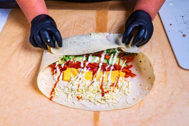 Siyah eldivenli biri, içi taze sebze, peynir ve sosla dolu bir ambalajı ustalıkla katlıyor. Hazırlık, gün ışığında temiz bir mutfak ortamında gerçekleştirilecek..