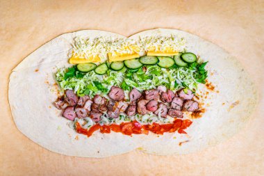 Büyük bir yassı ekmek cömertçe et, peynir, taze sebze ve canlı yeşilliklerle doludur. Yemek hazırlamada çeşitli doku ve renkler sergileniyor..