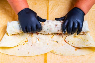 Siyah eldiven giyen eller lezzetli malzemelerle dolu yeni yapılmış bir sargı sarar. Mutfak düzeneği tortillaları dikkatlice yerleştirilmiş, canlı bir pişirme süreci sergiliyor..