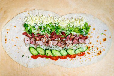 Yumuşak bir tortilla üzerinde et, taze marul, peynir, salatalık ve domates katmanları ile canlı bir paket hazırlanıyor. Her bileşen renkli ve lezzetli bir kombinasyon için düzenlenmiştir..