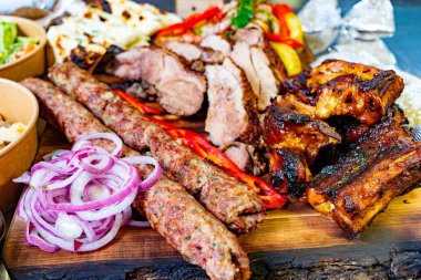Canlı bir tabakta, dilimlenmiş soğan ve renkli sebzelerle süslenmiş sosis, pirzola ve tavuk da dahil olmak üzere çeşitli ızgara etler sergileniyor. Yaz ziyafeti için mükemmel..