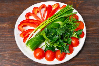 Parlak renkli sebzeler yuvarlak bir tabakta sanatsal olarak düzenlenir. Tabakta kırmızı çan biberi dilimleri, sulu domatesler, salatalık, yeşil soğan ve sağlıklı beslenmenin sergilendiği taze otlar yer alıyor..