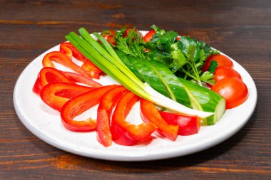 Parlak kırmızı çan biberleri, kiraz domatesleri, yeşil soğanlar ve taze bitkiler, beyaz bir tabakta, parlak renkler ve dokuların ön planda olduğu, koyu ahşap bir zemine karşı sanatsal olarak düzenlenmiştir..