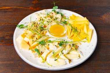 Çok güzel düzenlenmiş bir peynir tabağında çeşitli peynirler, bal sosu ve taze süsler bulunur. Sunum, rustik bir ahşap masadaki dokuları ve renkleri vurgular.