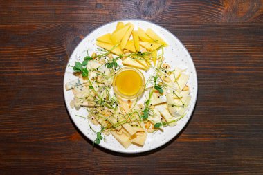 Çok güzel düzenlenmiş bir peynir tabağında çeşitli peynirlerin yanı sıra taze meyve dilimleri ve küçük bir bal tabağı yer alır. Yeşil otlar ortama tazelik katar..