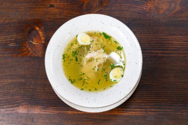 Taze otlar ve ince dilimlenmiş sebzelerle süslenmiş sıcak bir kase tavuk çorbası ahşap bir masada duruyor. Serin bir gün için harika bir yemek..