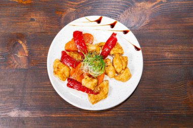Kırmızı biber ve taze sebzelerle harmanlanmış gevrek tavuk parçaları içeren canlı bir yemek. Sunumda kırsal bir ahşap masa üzerinde bir sos yağmuru ve yeşil garnitür yer alıyor..