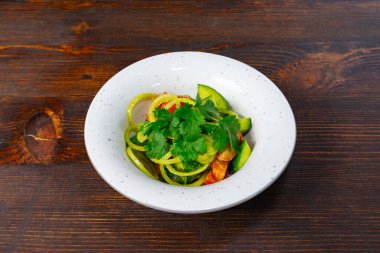 Dilimlenmiş salatalık, domates ve taze kişniş soslu soğan içeren canlı bir salata. Tabak, kırsal bir tahta masada beyaz bir kasede sunuluyor, renklerini sergiliyor..