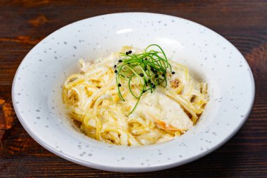 Narin bitkilerle servis edilen bir tabak kremalı makarna ve bir tutam baharat, ahşap bir masada güzelce sunuluyor. Yemek davetkar ve lezzetli görünüyor..