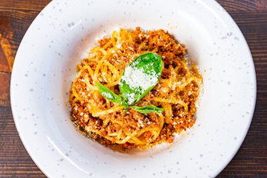 Bir tabak spagetti, üzerinde rendelenmiş peynir ve taze fesleğen yaprağı olan zengin bir et sosuyla servis edilir. Tabak kırsal ahşap bir masada sunuluyor..