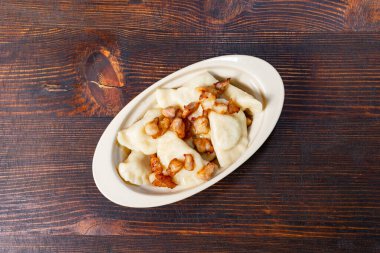 Lezzetli hamur köfteleri beyaz bir tabakta servis edilir, üzerinde bol bol kıtır pastırma parçaları vardır. Ahşap masa bu iştah açıcı yemeğe kırsal bir dokunuş katıyor..