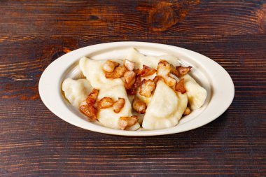 Patates ve peynirle dolu bir tabak yumuşak pierogi. Üstünde de altın kıtır pastırma parçaları var. Tabak kırsal ahşap bir masada sunuluyor, doyurucu ve geleneksel bir yemek sergileniyor..