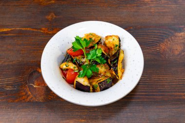 Patlıcan, domates ve otlar dahil olmak üzere canlı bir sebze karışımı, kırsal bir ahşap masada duran beyaz bir kasede güzelce düzenlenmiştir..