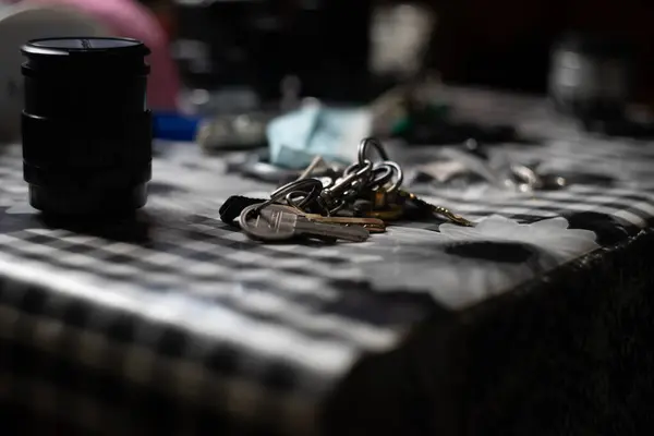 Anahtarlar kareli bir masa örtüsünün üzerinde dağınık eşyalar arasında duruyor. Yakınlarda oturan bir kamera merceği, yumuşak aydınlatılmış bir odada çeşitli objelerle çevrili, rahat bir atmosfer yaratıyor..