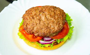 Taze marul, domates ve kırmızı soğanla kaplı, doyurucu bir sığır burger ekmeğin üzerine oturur. Yemek canlı renkleri vurguluyor ve sıradan bir yemek ortamında eğlenmeye hazır görünüyor..