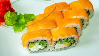 Taze suşi ruloları güzel bir şekilde beyaz bir tabakta sergileniyor. İçinde canlı portakallı somon balığı, kremalı ve çıtır çıtır sebzeler ve wasabi ve yeşillikler var..