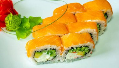Taze suşi ruloları güzel bir şekilde beyaz bir tabakta sergileniyor. İçinde canlı portakallı somon balığı, kremalı ve çıtır çıtır sebzeler ve wasabi ve yeşillikler var..